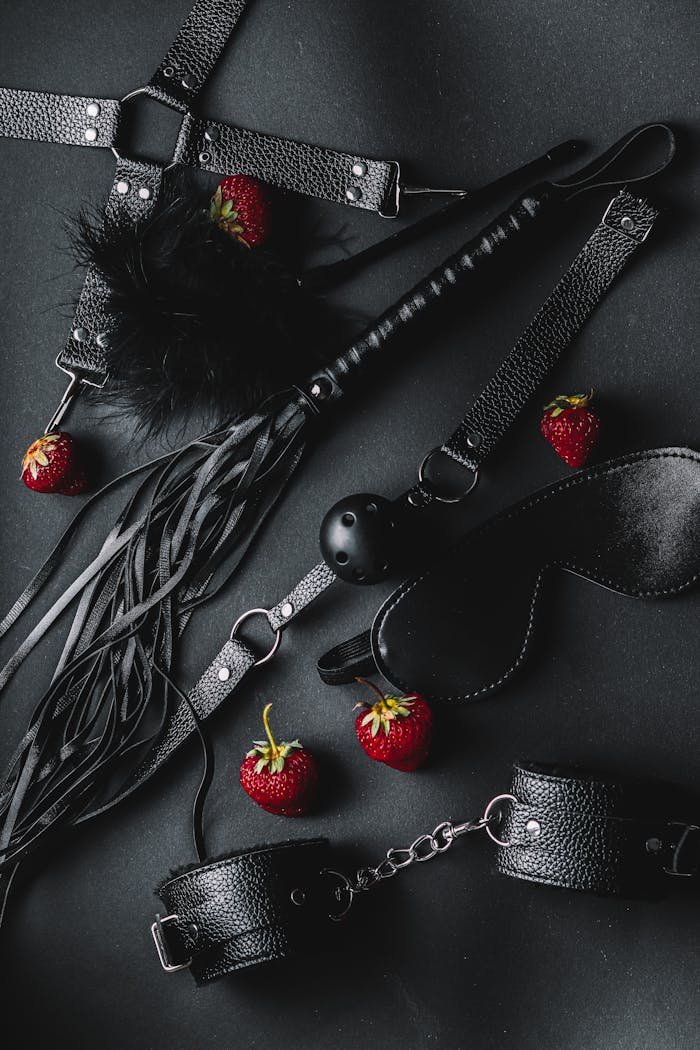 A stylish flat lay of leather BDSM accessories with fresh strawberries on a dark background.